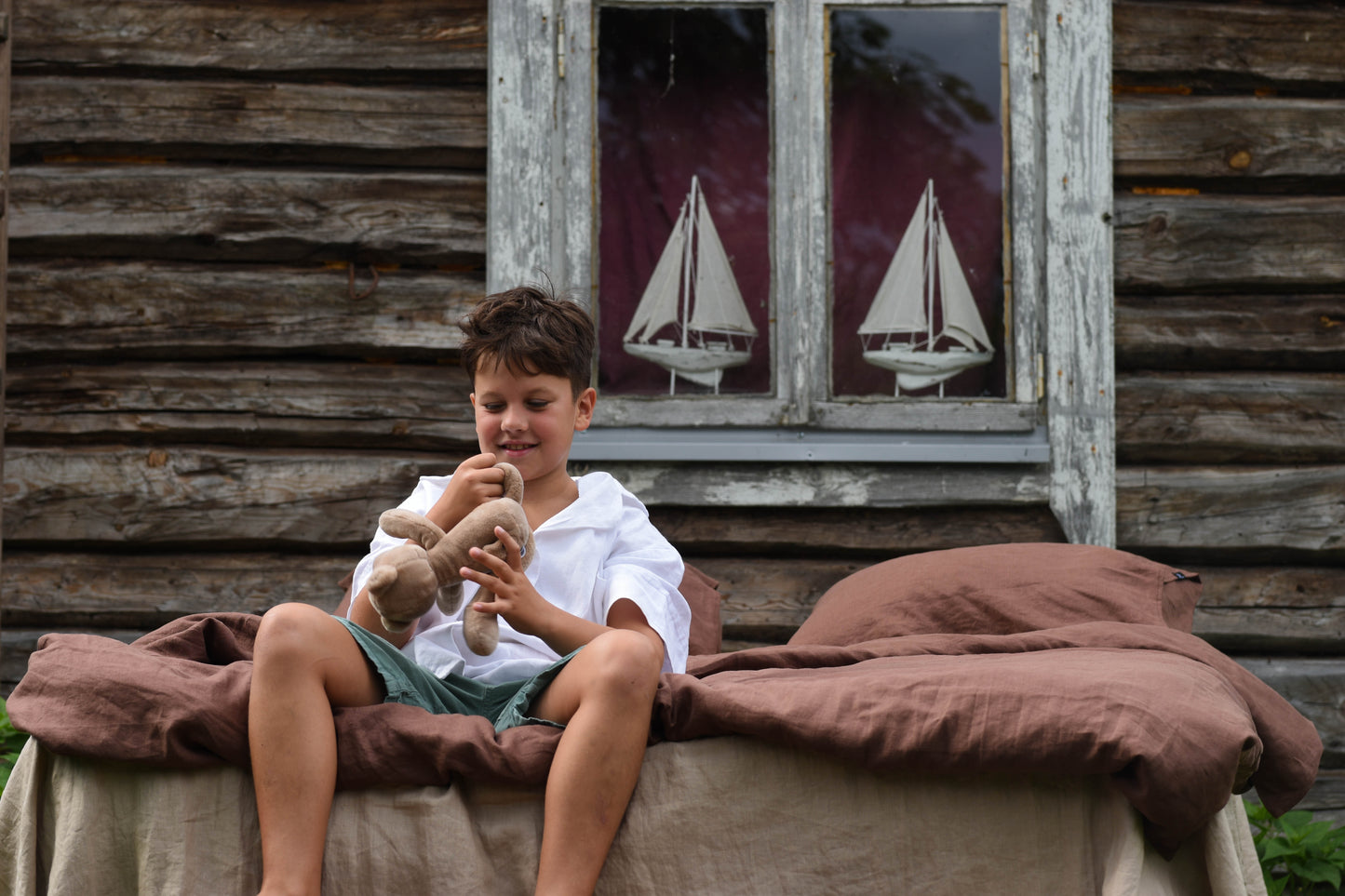 Young boy holding a teddy bear in front of a wooden cabin with sailboat decorations.