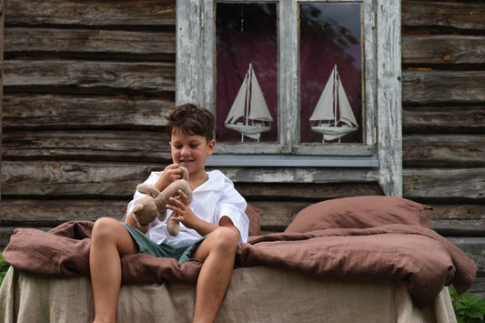Young boy holding a teddy bear in front of a wooden cabin with sailboat decorations.