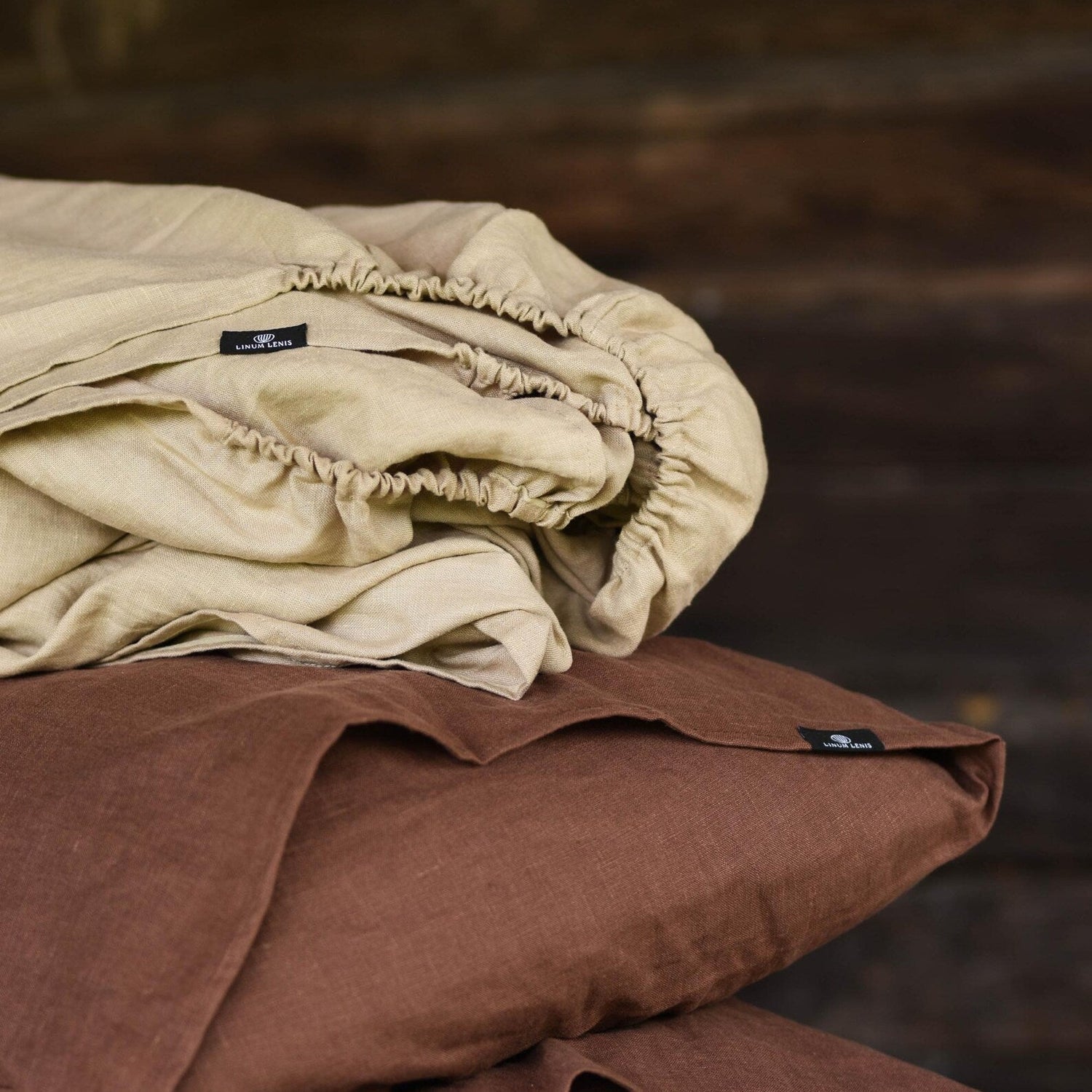 Organic linen fitted sheet in warm sand beige stacked on chocolate brown linen pillowcases, soft stonewashed texture, minimalist bedroom aesthetic.