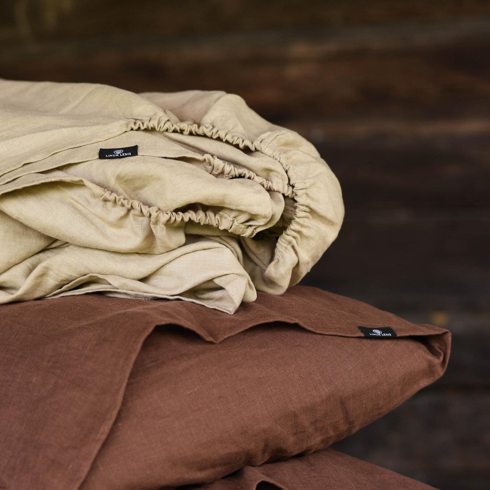 Organic linen fitted sheet in warm sand beige stacked on chocolate brown linen pillowcases, soft stonewashed texture, minimalist bedroom aesthetic.
