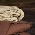 Organic linen fitted sheet in warm sand beige stacked on chocolate brown linen pillowcases, soft stonewashed texture, minimalist bedroom aesthetic.
